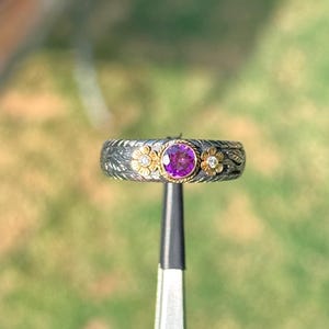 May include: A silver ring featuring a central round purple gemstone set in a gold bezel. The band has a dark, textured finish with engraved leaf patterns and two small gold flower accents, each containing a tiny clear stone on either side.