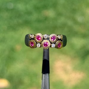 May include: A detailed shot of a ring featuring a dark band with alternating pink gemstones and small clear stones set in gold-colored bezels. The ring is held by a tool against a blurred green background.