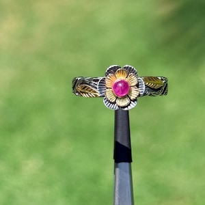 May include: A silver ring with a flower design. The flower has a gold center with a pink gemstone and gold petals. The band has a leaf pattern with gold accents. The ring is held by a black tool against a green background.