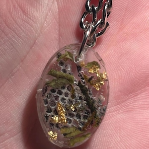 May include: Oval pendant necklace with a silver chain. The clear resin pendant contains dried leaves, small black seeds, and gold flakes. The pendant is approximately 1 inch long.