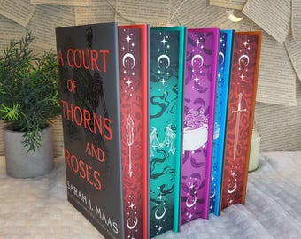 May include: A collection of hardcover books with colorful spines. The book covers feature the title "A Court of Thorns and Roses" in red text. The spines are decorated with various illustrations and patterns, including moons, stars, and a sword.