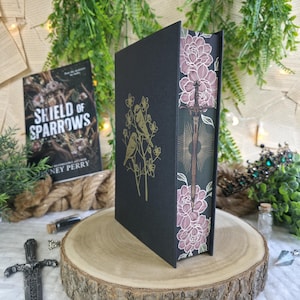 Op de afbeelding: Een hardcover boek getiteld "Shield of Sparrows" met een zwarte kaft met gouden vogelillustraties. De rug toont een zwaardontwerp en bloemmotieven in roze en groen. Het boek wordt gepresenteerd op een houten schijf.