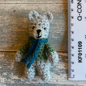 May include: A small crocheted teddy bear wearing a green and blue scarf. The bear is about 2 inches tall and is sitting next to a ruler.