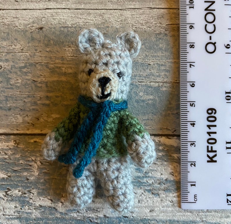 Pu&ograve; includere: Un piccolo orsacchiotto di peluche all'uncinetto con una sciarpa verde e blu. L'orsacchiotto &egrave; alto circa 5 cm ed &egrave; posizionato accanto a un righello.