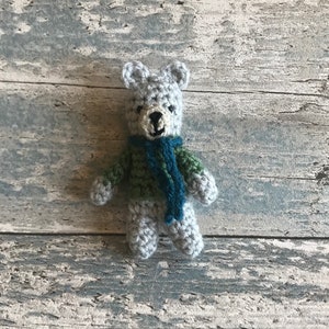 Pu&ograve; includere: Un piccolo orsacchiotto di peluche grigio all'uncinetto con una sciarpa verde e blu. L'orsacchiotto &egrave; seduto su una superficie di legno grigio chiaro.