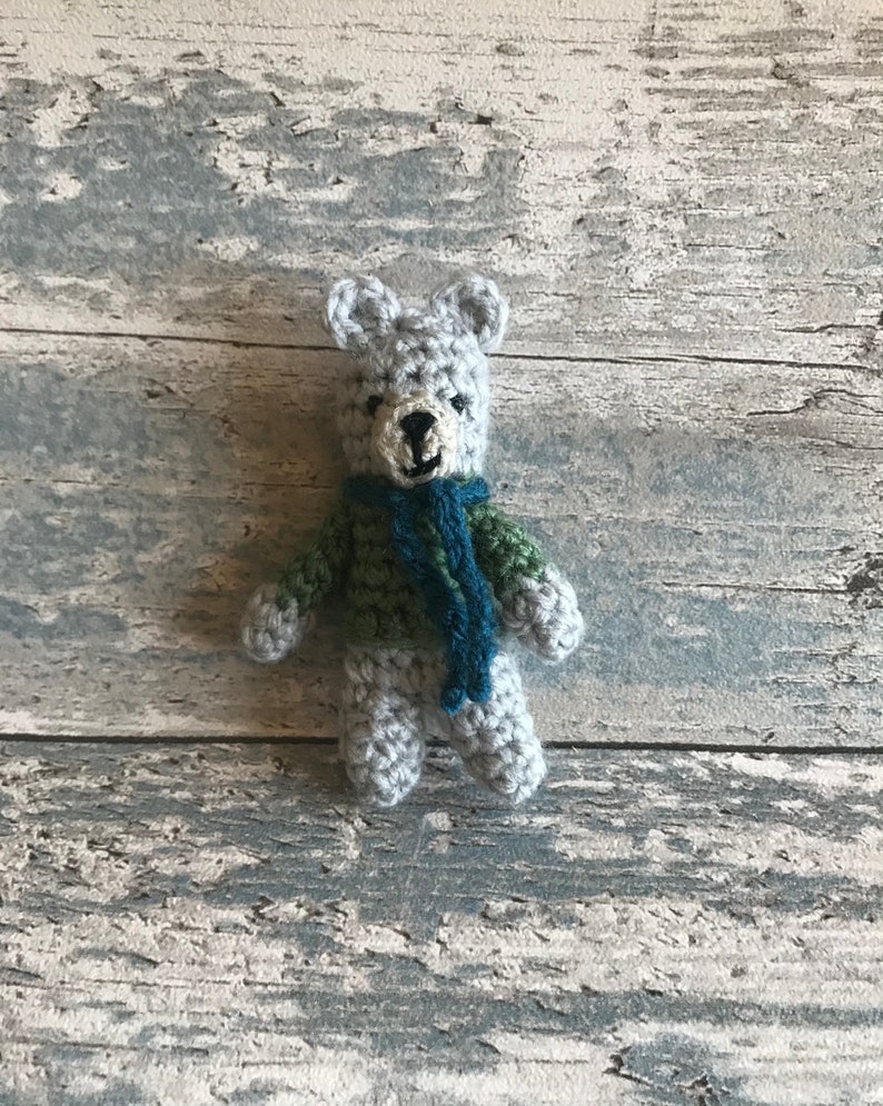 Pu&ograve; includere: Un piccolo orsacchiotto di peluche grigio all'uncinetto con una sciarpa verde e blu. L'orsacchiotto &egrave; seduto su una superficie di legno grigio chiaro.