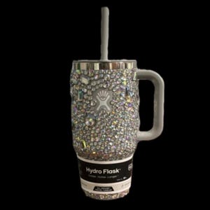 May include: A silver Hydro Flask tumbler with a clear lid and a straw. The tumbler is covered in iridescent rhinestones. The text "Hydro Flask" is printed on the bottom of the tumbler.