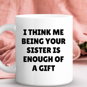 May include: A white ceramic mug featuring the text "I THINK ME BEING YOUR SISTER IS ENOUGH OF A GIFT" in bold black lettering. The mug has a curved handle and is set against a soft pink background with floral accents.