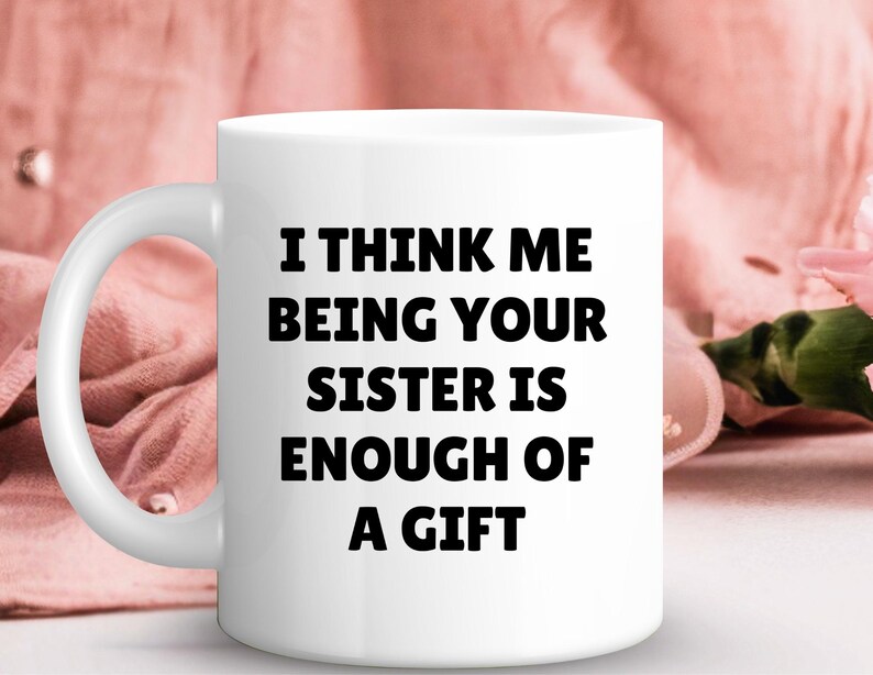 May include: A white ceramic mug featuring the text "I THINK ME BEING YOUR SISTER IS ENOUGH OF A GIFT" in bold black lettering. The mug has a curved handle and is set against a soft pink background with floral accents.