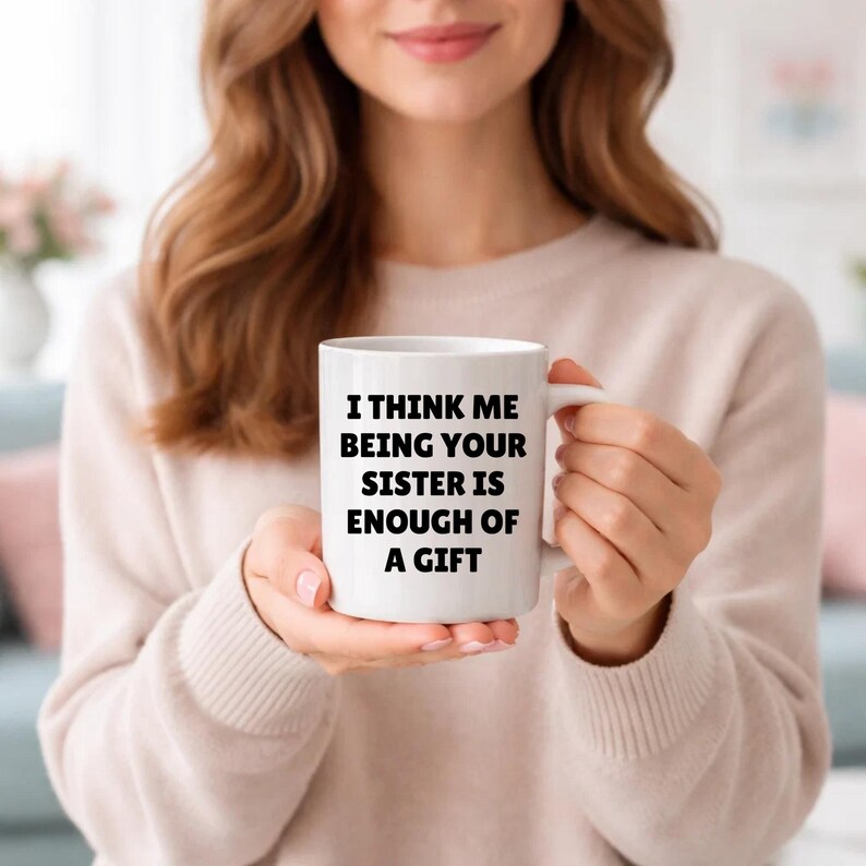 May include: White ceramic mug with the text "I THINK ME BEING YOUR SISTER IS ENOUGH OF A GIFT" in black. The mug has a white handle and is held by a person wearing a light-coloured jumper.