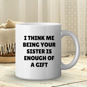 May include: White ceramic mug with a curved handle. The mug features the text "I THINK ME BEING YOUR SISTER IS ENOUGH OF A GIFT" in black, centered on the front. The mug is on a patterned rug.