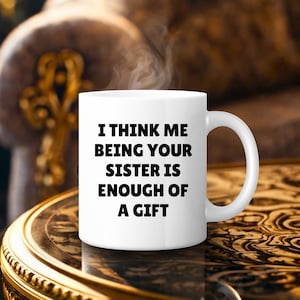 May include: A white ceramic mug with the text "I THINK ME BEING YOUR SISTER IS ENOUGH OF A GIFT" in black. The mug has a curved handle and is filled with a hot beverage, with steam rising from the top. The mug is on a decorative table.