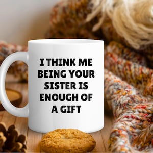 May include: White ceramic mug with the text "I THINK ME BEING YOUR SISTER IS ENOUGH OF A GIFT" in black. The mug is on a wooden surface with a biscuit and a decorative scarf in the background.