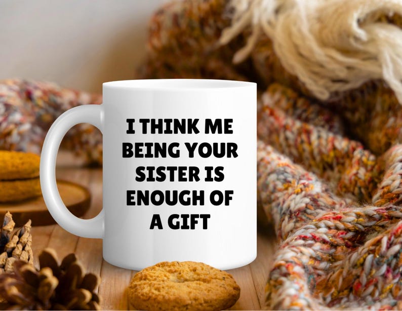 May include: White ceramic mug with the text "I THINK ME BEING YOUR SISTER IS ENOUGH OF A GIFT" in black. The mug is on a wooden surface with a biscuit and a decorative scarf in the background.