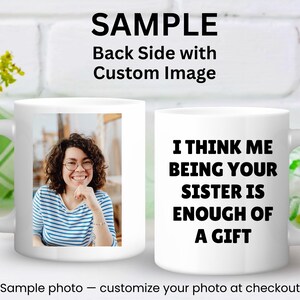 May include: White ceramic mug featuring a photo of a smiling person on one side and the text "I THINK ME BEING YOUR SISTER IS ENOUGH OF A GIFT" on the other. The mug has a curved handle and is set against a white brick background.