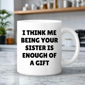 May include: White ceramic mug with a curved handle. The mug features the black text: "I THINK ME BEING YOUR SISTER IS ENOUGH OF A GIFT". The mug is on a marble surface.