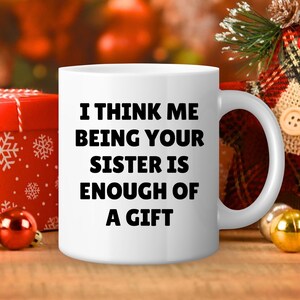 May include: White ceramic mug with the text "I THINK ME BEING YOUR SISTER IS ENOUGH OF A GIFT" in black. The mug is on a wooden surface with Christmas decorations in the background.