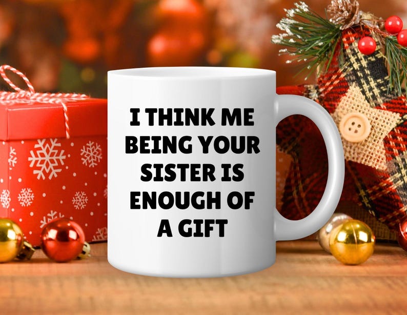 May include: White ceramic mug with the text "I THINK ME BEING YOUR SISTER IS ENOUGH OF A GIFT" in black. The mug is on a wooden surface with Christmas decorations in the background.