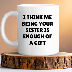 May include: White ceramic mug with the text "I THINK ME BEING YOUR SISTER IS ENOUGH OF A GIFT" in black. The mug is on a rustic wooden coaster. The background has a warm, blurred gradient.