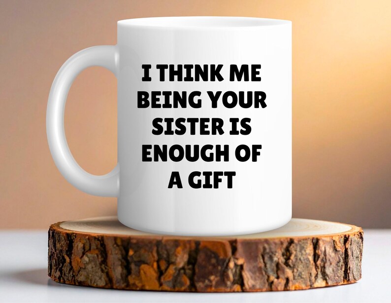 May include: White ceramic mug with the text "I THINK ME BEING YOUR SISTER IS ENOUGH OF A GIFT" in black. The mug is on a rustic wooden coaster. The background has a warm, blurred gradient.