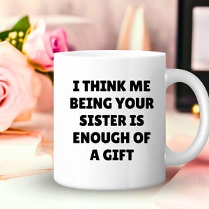 May include: White ceramic mug with the text "I THINK ME BEING YOUR SISTER IS ENOUGH OF A GIFT" in black. The mug has a curved handle and is set against a blurred background with pink roses and a book.