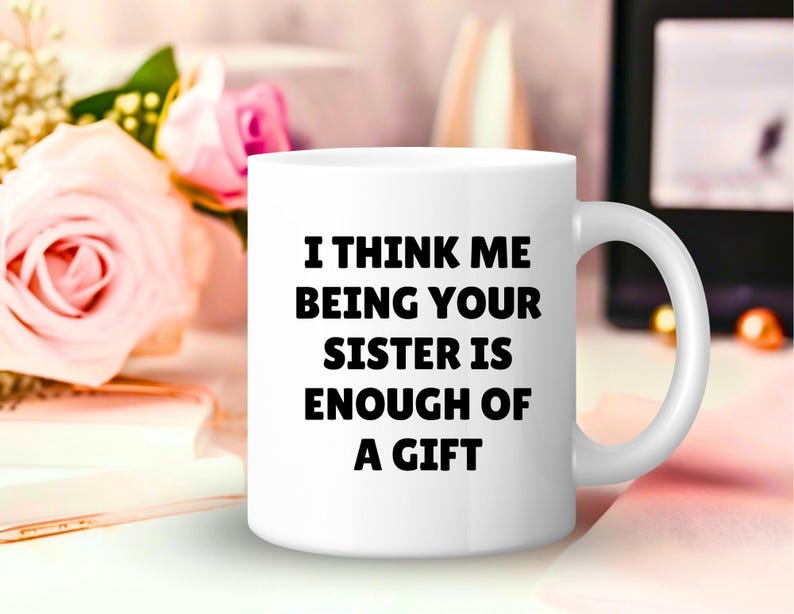 May include: White ceramic mug with the text "I THINK ME BEING YOUR SISTER IS ENOUGH OF A GIFT" in black. The mug has a curved handle and is set against a blurred background with pink roses and a book.