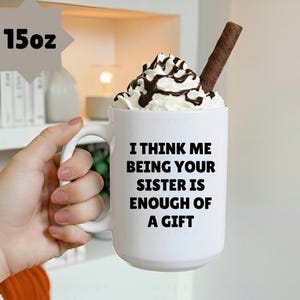May include: A white 15oz mug with the text "I THINK ME BEING YOUR SISTER IS ENOUGH OF A GIFT" in black. The mug is filled with whipped cream, chocolate syrup, and a chocolate wafer stick.