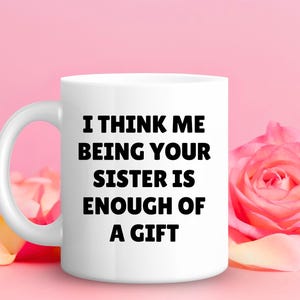 May include: White ceramic mug with the text "I THINK ME BEING YOUR SISTER IS ENOUGH OF A GIFT" in bold black letters. The mug is set against a pink background with a pink rose and petals.