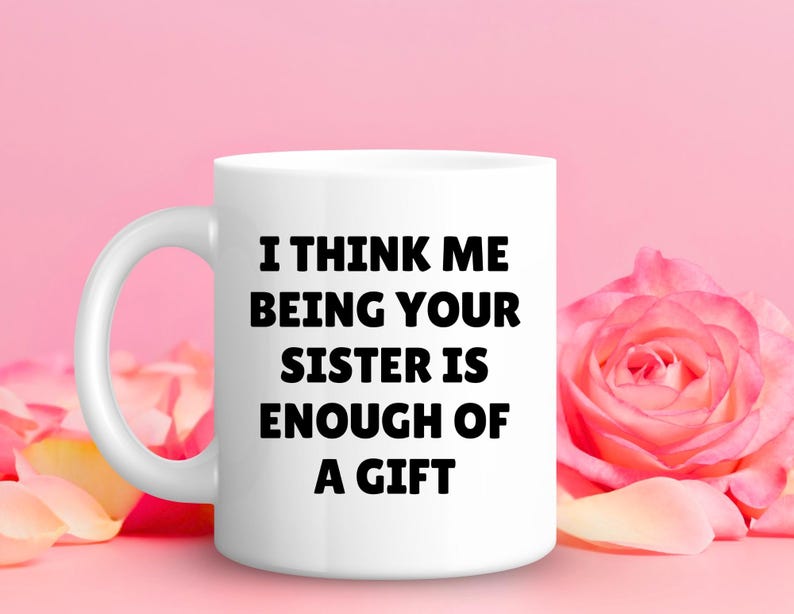 May include: White ceramic mug with the text "I THINK ME BEING YOUR SISTER IS ENOUGH OF A GIFT" in bold black letters. The mug is set against a pink background with a pink rose and petals.