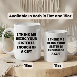 May include: Two white ceramic mugs with black text that reads "I THINK ME BEING YOUR SISTER IS ENOUGH OF A GIFT." The mugs are labeled 15oz and 11oz. The text "Available In Both in 11oz and 15oz" is above the mugs.