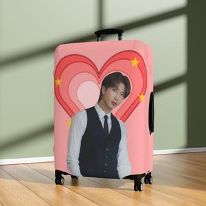 May include: Pink suitcase with a heart design and a yellow star. The heart has a layered design and a photo of a person with dark hair wearing a black waistcoat and white shirt. The suitcase has a black handle and wheels.