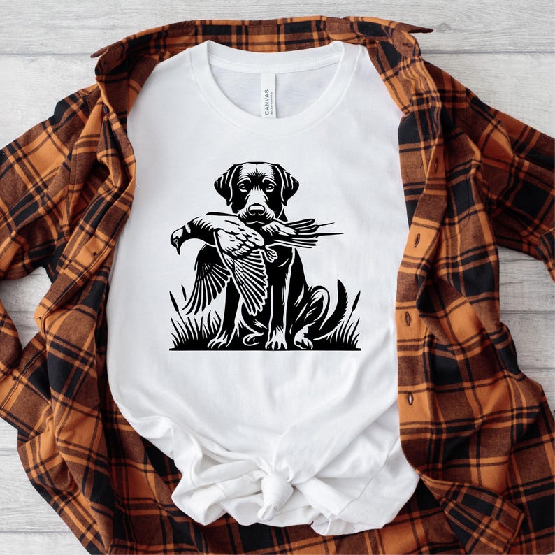 K&ouml;nnte beinhalten: Ein wei&szlig;es T-Shirt mit einer Schwarzwei&szlig;illustration eines schwarzen Labrador Retrievers, der einen Fasan im Maul h&auml;lt. Der Hund steht in einem grasbewachsenen Gebiet.