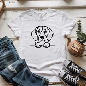 Puede incluir: Camiseta blanca con un dibujo lineal negro de un perro beagle. La cabeza y las patas del perro son visibles, dando la impresi&oacute;n de que est&aacute; mirando por encima de un borde. La camiseta se combina con vaqueros azules y zapatillas de lona negras.