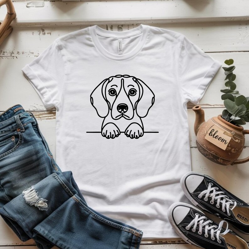 Puede incluir: Camiseta blanca con un dibujo lineal negro de un perro beagle. La cabeza y las patas del perro son visibles, dando la impresi&oacute;n de que est&aacute; mirando por encima de un borde. La camiseta se combina con vaqueros azules y zapatillas de lona negras.