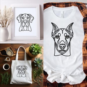 May include: A set of three white tote bags featuring black line drawings of dog breeds. The tote bags are displayed on a wooden surface alongside a white framed picture and a white t-shirt with a black line drawing of a dog breed.