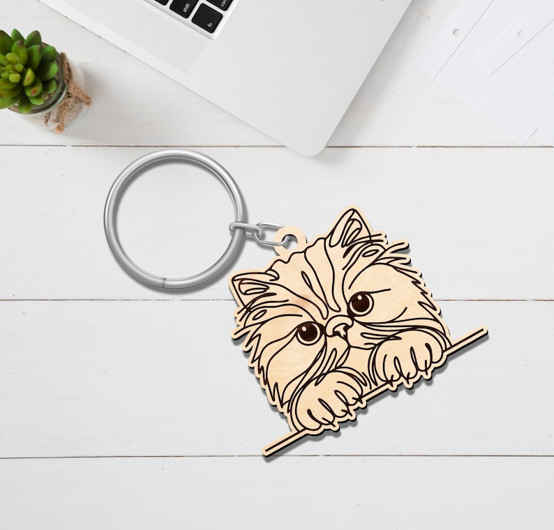 May include: A wooden keychain featuring a Persian cat design. The cat's face is intricately carved with detailed fur and large eyes. The keychain includes a silver-colored metal ring for attaching keys.