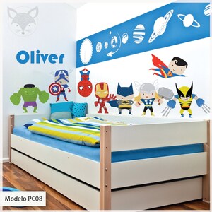 Puede incluir: Una cama con estructura blanca, con una manta a rayas azules y amarillas y una almohada azul y blanca. La pared detrás de la cama está decorada con un mural azul y blanco con planetas, estrellas y superhéroes. El nombre "Oliver" está escrito en letras negras en la pared.