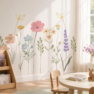 Op de afbeelding: Een muursticker set met aquarel-stijl wilde bloemen in tinten roze, blauw, geel en paars met groene stengels en bladeren. De bloemillustraties zijn aangebracht op een effen muur boven een houten boekenplank en een ronde tafel met stoelen.