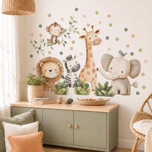 Op de afbeelding: Een set muurstickers met aquarelillustraties van een aap, leeuw, zebra, giraf en olifant, omringd door groene en beige stippen, met groene bladeren aan de voeten van de dieren afgebeeld in een zachte tekenstijl.