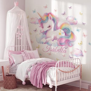 May include: A white metal bed with pink bedding, a woven basket, and a canopy with a crown. A wall decal features a unicorn with a rainbow mane and the name "Isabella". Butterflies decorate the wall, creating a whimsical bedroom theme.