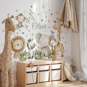 Puede incluir: Un conjunto de calcomanías de pared para guardería con una jirafa, un león, una cebra, un elefante y un mono. El arte mural incluye el nombre "Oliver" en escritura cursiva, con lunares verdes y beige. Un mueble de almacenamiento de madera con cubos blancos está en primer plano.