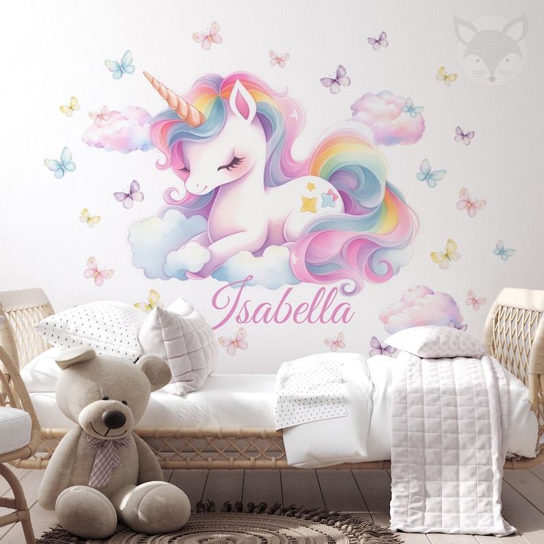 May include: A whimsical wall decal featuring a pastel-colored unicorn with a rainbow mane and tail, resting on fluffy clouds. Butterflies and the name "Isabella" in script surround the unicorn. The decal is placed above a bed with a teddy bear and pillows.