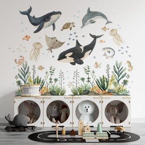 Puede incluir: Una escena caprichosa de guardería con una unidad de almacenamiento blanca con compartimentos redondos, cada uno con un juguete de peluche. Arriba, un mural de pared representa varias criaturas marinas como ballenas, delfines y medusas. Una alfombra de juego negra con un diseño de carretera y juguetes de madera completan la escena.