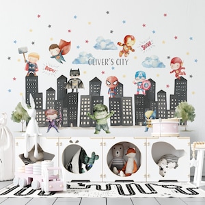 Puede incluir: Un vinilo decorativo blanco con personajes de superhéroes y un diseño de paisaje urbano. El diseño incluye ilustraciones de superhéroes, edificios, nubes y estrellas. El texto "OLIVER'S CITY" también está incluido. Un mueble de almacenamiento blanco con figuras de juguete está en primer plano.