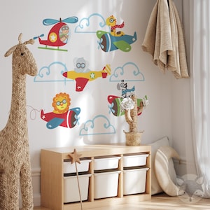May include: A set of colorful animal-themed wall decals featuring a giraffe, a lion, a zebra, a koala, and a bird in airplanes and a helicopter. The decals are on a white wall with clouds and are perfect for a child's room.