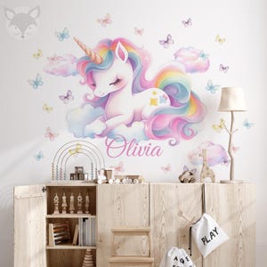 May include: A whimsical wall decal featuring a pastel unicorn with a rainbow mane and horn, resting on a cloud. The name "Olivia" is written below the unicorn. Butterflies and clouds decorate the white wall. A wooden cabinet with toys is in the foreground.