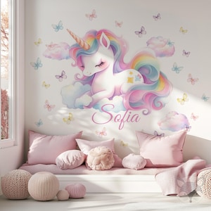 May include: A pastel unicorn wall sticker with a rainbow mane and horn, resting on a cloud. The name "Sofia" is written below. Butterflies and clouds decorate the wall. Pink cushions and pouffes complete the dreamy scene.