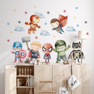 Op de afbeelding: Een set muurstickers in aquarel-stijl met chibi-geïnspireerde superheldenfiguren zoals Iron Man, Superman, Captain America, Thor, Spider-Man, Hulk en Batman, vergezeld van verspreide blauwe, rode, gele en grijze sterren en wolken.