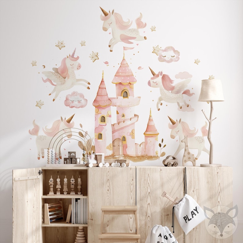 May include: Pink and gold unicorn wall decals with a pink castle and clouds. The decals are on a white wall above a wooden cabinet with shelves.
