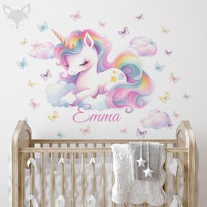 May include: A whimsical wall decal featuring a pastel unicorn with a rainbow mane and tail, resting on fluffy clouds. Butterflies in various colors surround the unicorn. The name "Emma" is written in cursive below the unicorn.